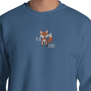Pu&ograve; includere: Una felpa blu con una piccola volpe ricamata e il testo "FOR FOX SAKE" in bianco.