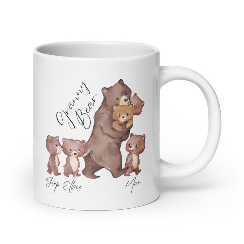 May include: White ceramic mug with a brown bear illustration. The bear is holding two smaller bears. The text "Granny Bear" is written in cursive above the bear. The text "Shep Elfie Max" is written below the bear.