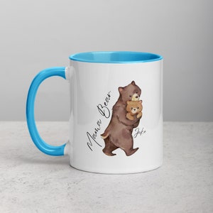 May include: White ceramic mug with a light blue rim and handle. The mug features a watercolor illustration of a brown bear holding a smaller brown bear cub. The text "Mama Bear" is written in cursive on the left side of the mug and the text "Jake" is written on the right side of the mug.