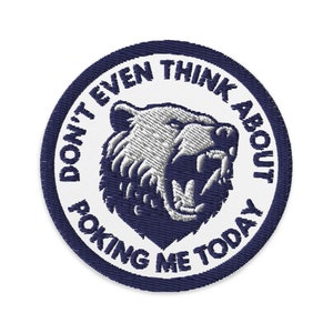 Può includere: Un cerotto rotondo bianco con un bordo blu scuro. Il cerotto presenta un orso bianco con una bocca blu e un contorno blu scuro. Il testo "Don't even think about poking me today" è scritto in un cerchio attorno all'orso.