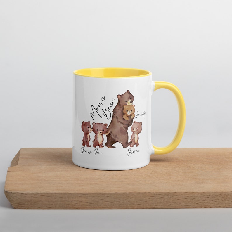 May include: White ceramic mug with a yellow rim. The mug features a watercolor illustration of a brown bear with three cubs. The text "Mama Bear" is written in cursive above the bears. The cubs are labeled "James", "Joan", and "Jessica".