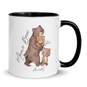 May include: White ceramic mug with a black rim and handle. The mug features a watercolor illustration of a brown bear holding a smaller brown bear cub. The text "Mama Bear" is written vertically on the left side of the mug. The text "Jake" is written on the right side of the mug. The text "Annabel" is written below the bears.