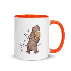 May include: White ceramic mug with an orange rim and handle. The mug features a watercolor illustration of a brown bear holding a smaller brown bear cub. The text "Mama Bear" is written vertically on the left side of the mug and the text "Jake" is written below the bears.