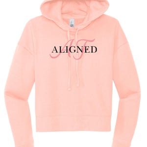 May include: A light peach-colored cropped hoodie with a drawstring hood. The word "ALIGNED" is printed across the chest in black and pink script. The hoodie has long sleeves and a ribbed hem and cuffs. A casual, comfortable top.