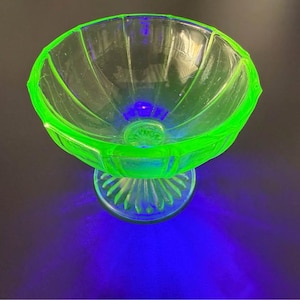 May include: A translucent, vibrant neon green glass bowl with a fluted, scalloped rim and a pedestal base. The glass exhibits a bright, glowing appearance under ultraviolet light, featuring vertical ridges along the bowl and the circular base.