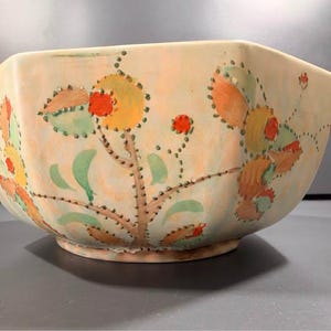 May include: A large, cream-coloured ceramic bowl with a faceted, multi-sided shape. The exterior features a hand-painted botanical design with stylised flowers and leaves in shades of orange, yellow, and green, accented by small, raised dark dots.