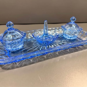 May include: A light blue glass vanity set consisting of a rectangular tray with a sunburst pattern, two small lidded jars, and a central ring holder. The glass features a pressed geometric design along the edges and a translucent finish.