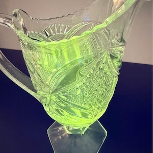 May include: A translucent lime green glass jug featuring a pressed pattern of geometric diamonds, starbursts, and fan motifs. The vessel has a scalloped rim, a curved handle, and a hexagonal base, exhibiting a vibrant glow characteristic of uranium glass.