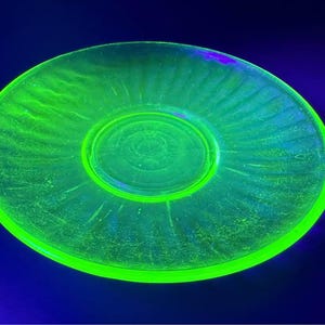 May include: A circular glass saucer glowing with a vibrant neon green hue under ultraviolet light. The saucer features a smooth, raised central circular area surrounded by a wide, flat rim decorated with a series of radiating, linear indentations.