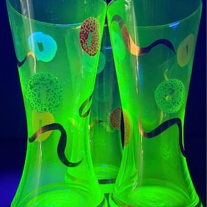 May include: Three translucent green glass vases glowing under ultraviolet light. Each vase features a flared rim and a tapered body decorated with scattered circular patterns in red, yellow, and blue, accented by thin, dark, wavy black lines.