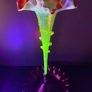 May include: A tall, slender glass vase with a vibrant, glowing lime green stem and a wide, ruffled rim featuring a deep red edge. The stem is decorated with a spiralling glass coil, and the entire piece exhibits a bright, fluorescent appearance.