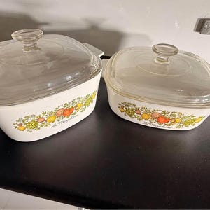 May include: Two white rectangular casserole dishes with clear glass lids. Each dish features a colourful vegetable pattern including tomatoes, lemons, and artichokes, with the script text La Marjolaine printed on the front of each white ceramic base.