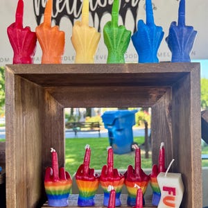 May include: A collection of wax candles shaped like hands making a middle finger gesture. The top row features solid red, orange, yellow, green, blue, and purple candles. The bottom row includes rainbow-colored hand candles and candles with the text FUCK IT.