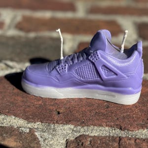 May include: A lavender-colored sneaker-shaped candle with white accents on the sole. The candle features intricate details mimicking a popular athletic shoe design, with two wicks extending from the top.