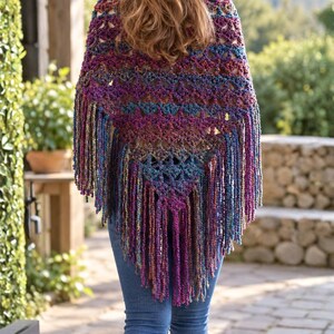 May include: A triangular crochet shawl featuring a variegated yarn pattern in shades of purple, blue, and gold. The garment has an openwork stitch design and is finished with long, thick fringe tassels along the bottom edges and the pointed hem.