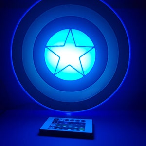 Puede incluir: Una luz nocturna con forma de escudo del Capitán América azul y blanco con una estrella blanca en el centro. La luz está encendida y brilla en azul. Un mando a distancia negro con botones está en primer plano.