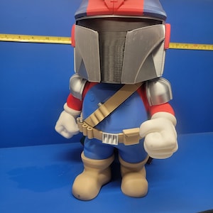 Pode incluir: Uma figura impressa em 3D de um personagem vestindo um macac&atilde;o azul, um cinto marrom com uma bolsa e um capacete vermelho e azul com um "M" branco na frente. O personagem tem luvas brancas e botas de cor bege.