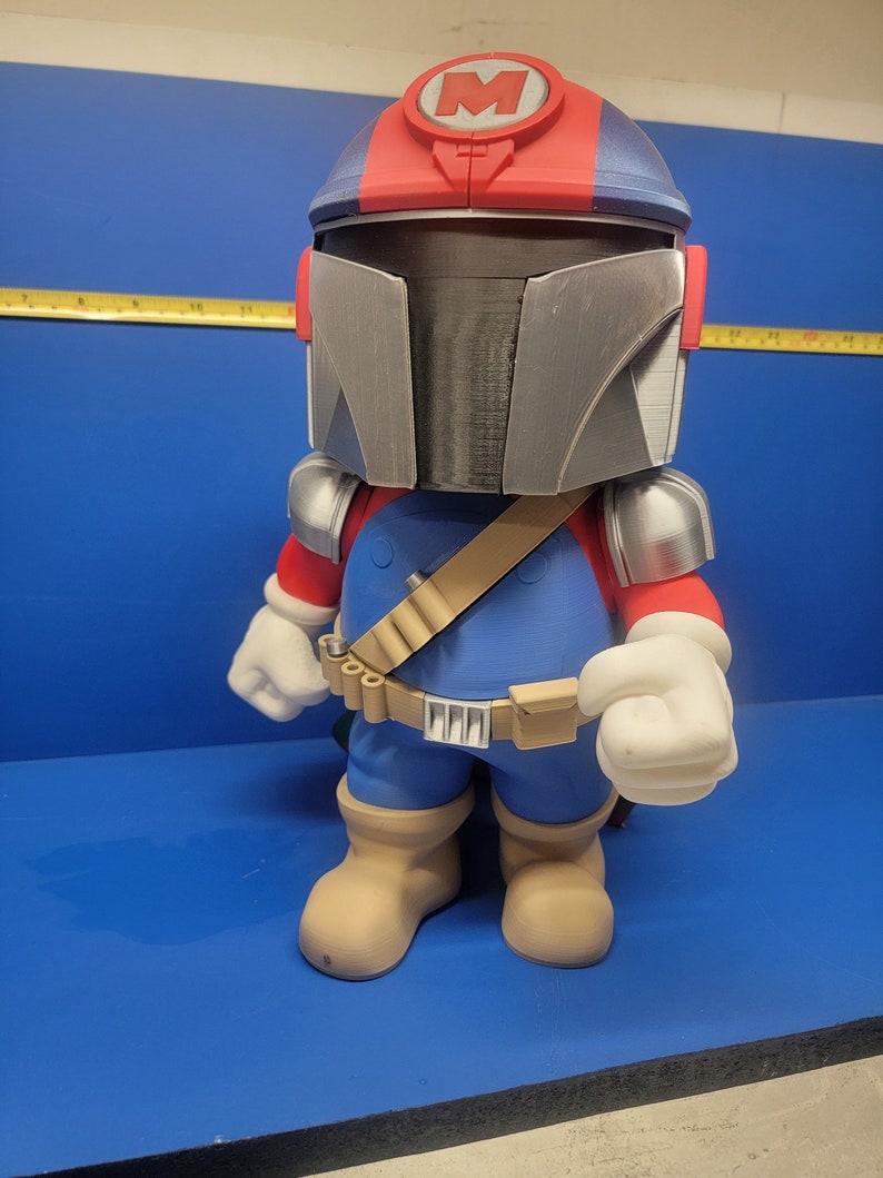 Pode incluir: Uma figura impressa em 3D de um personagem vestindo um macac&atilde;o azul, um cinto marrom com uma bolsa e um capacete vermelho e azul com um "M" branco na frente. O personagem tem luvas brancas e botas de cor bege.
