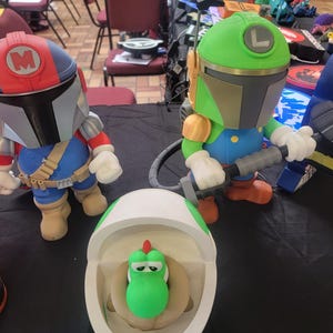 Pode incluir: Tr&ecirc;s figuras impressas em 3D, uma &eacute; um Mandaloriano verde e dourado com uma arma preta, outra &eacute; um Mandaloriano vermelho e azul com um 'M' branco no capacete, e a &uacute;ltima &eacute; um Yoshi verde em um vaso sanit&aacute;rio branco.