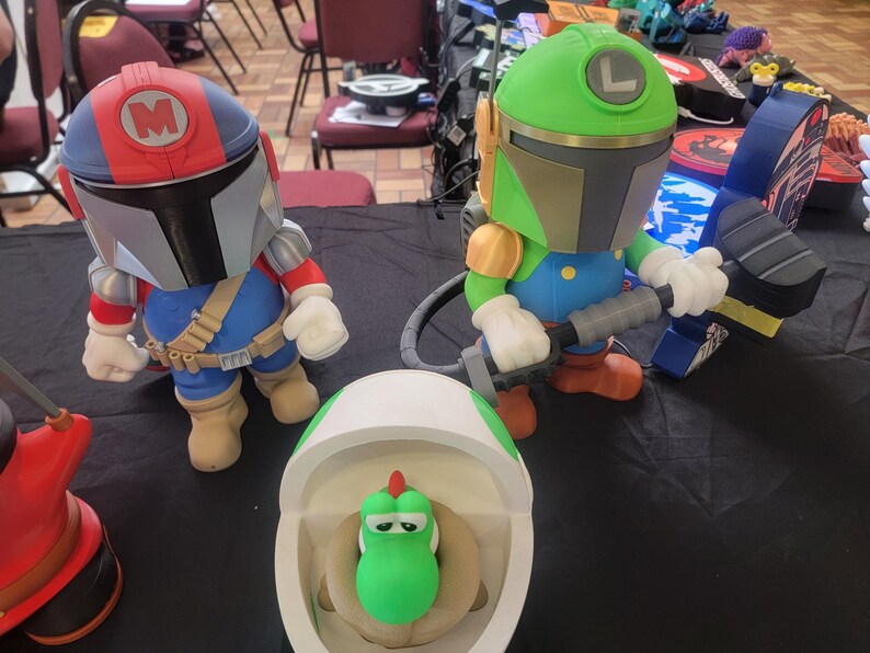 Pode incluir: Tr&ecirc;s figuras impressas em 3D, uma &eacute; um Mandaloriano verde e dourado com uma arma preta, outra &eacute; um Mandaloriano vermelho e azul com um 'M' branco no capacete, e a &uacute;ltima &eacute; um Yoshi verde em um vaso sanit&aacute;rio branco.