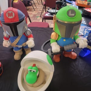 Pode incluir: Tr&ecirc;s figuras de brinquedo, uma figura de estilo mandaloriano vermelha e azul com um 'M' no capacete, uma figura verde e azul com um 'L' no capacete, e uma figura verde Yoshi dentro de um ovo branco.