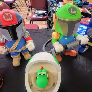 Pode incluir: Tr&ecirc;s figuras de brinquedo, uma figura verde e branca com um capacete, uma figura vermelha e azul com um capacete e uma figura verde com uma concha branca. As figuras est&atilde;o sobre uma superf&iacute;cie preta.