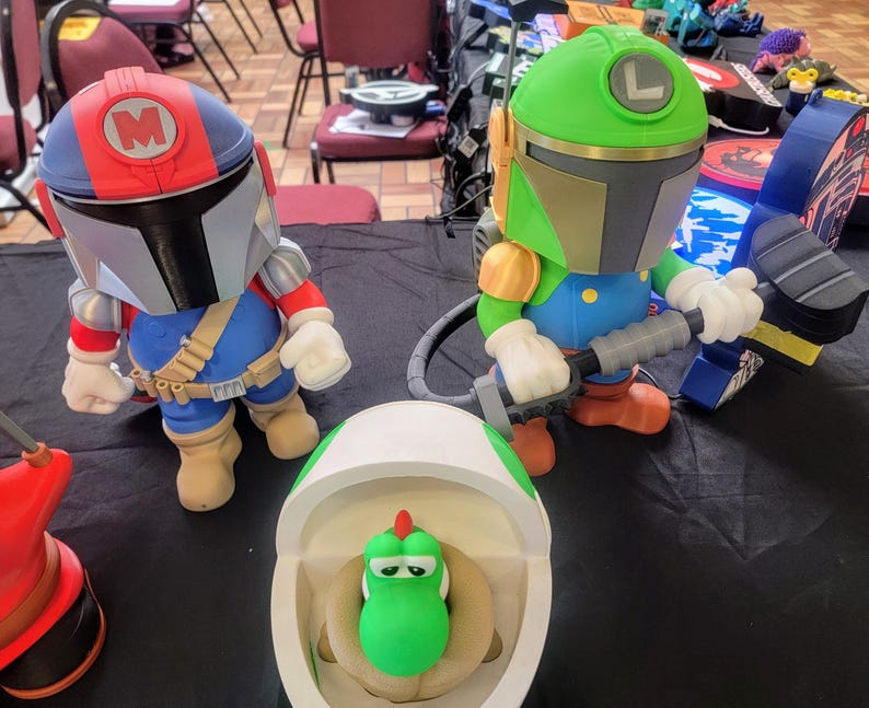 Pode incluir: Tr&ecirc;s figuras de brinquedo, uma figura verde e branca com um capacete, uma figura vermelha e azul com um capacete e uma figura verde com uma concha branca. As figuras est&atilde;o sobre uma superf&iacute;cie preta.
