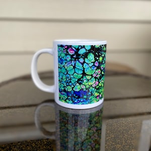 Könnte beinhalten: Weiße Keramiktasse mit einem farbenfrohen abstrakten Design in Blau, Grün und Rosa. Das Design ähnelt einem Mosaik oder einer Buntglasscheibe.