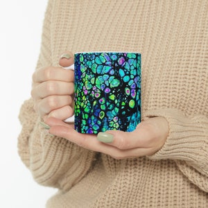 May include: A white ceramic mug with a colourful abstract design. The design features a swirling pattern of green, blue, and purple colours.