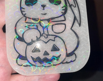 May include: A rectangular, iridescent sticker featuring a cartoon cat holding a knife and a jack-o'-lantern. The cat is white with black outlines and large eyes. The jack-o'-lantern has a carved face. The sticker has a holographic effect.