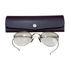 May include: Antique eyeglasses with a gold-tone frame and oval lenses, resting in front of a dark brown leather case. The case has a silver-tone button closure. The glasses have wire temples.