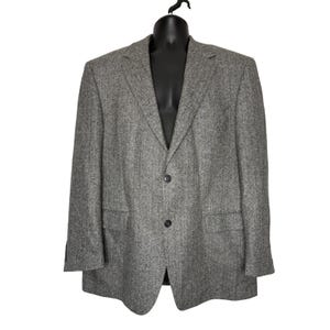 May include: A grey blazer featuring a subtle herringbone pattern, a notched lapel, and a two-button front closure. The garment includes two flap pockets at the waist and long sleeves with buttoned cuffs, presented on a black mannequin.