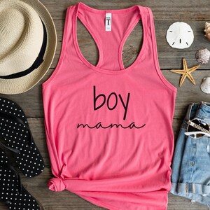 Puede incluir: Una camiseta sin mangas rosa con las palabras "boy mama" en escritura negra. La camiseta se muestra sobre una superficie de madera con un sombrero de paja, chanclas, pantalones cortos de mezclilla, gafas de sol, conchas marinas y una estrella de mar.