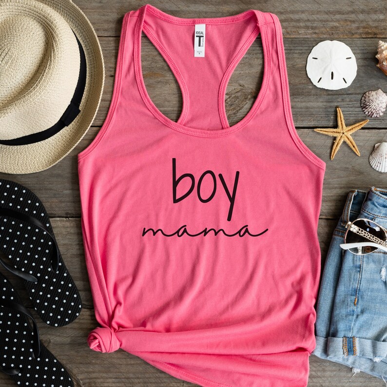 Puede incluir: Una camiseta sin mangas rosa con las palabras "boy mama" en escritura negra. La camiseta se muestra sobre una superficie de madera con un sombrero de paja, chanclas, pantalones cortos de mezclilla, gafas de sol, conchas marinas y una estrella de mar.