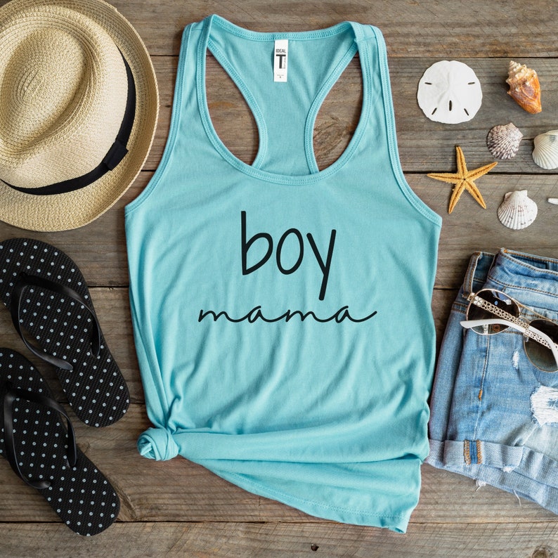 Puede incluir: Una camiseta sin mangas azul claro con la frase "boy mama" en negro. La imagen incluye un sombrero de paja, chanclas de lunares blancos y negros, pantalones cortos de mezclilla, gafas de sol y conchas marinas sobre una superficie de madera.