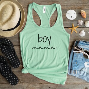 Puede incluir: Una camiseta sin mangas verde menta con las palabras "boy mama" en escritura negra. La camiseta est&aacute; sobre una superficie de madera con un sombrero de paja, chanclas, pantalones cortos de mezclilla, gafas de sol y conchas marinas.