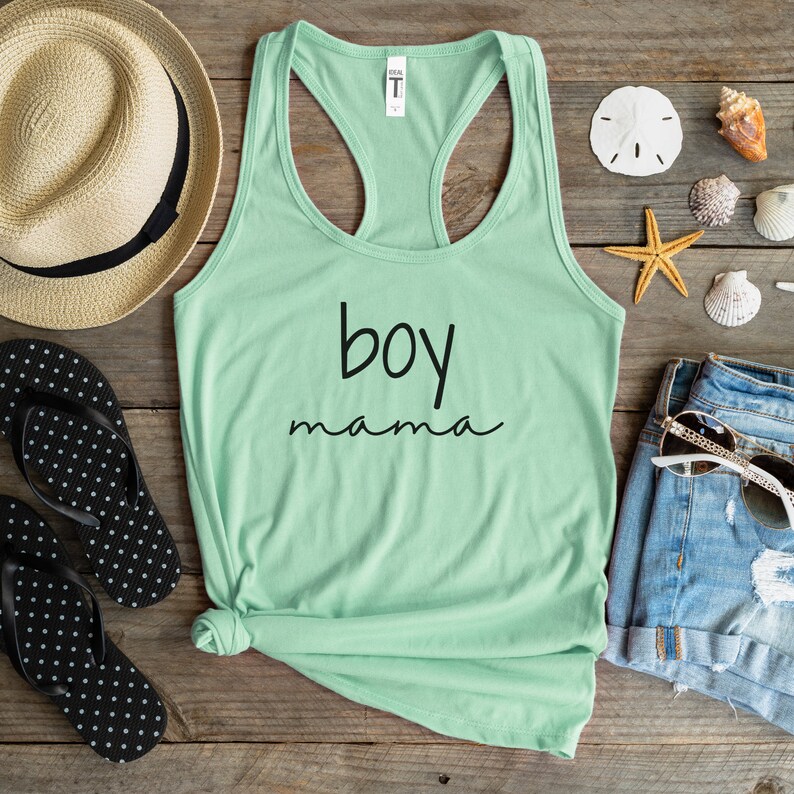 Puede incluir: Una camiseta sin mangas verde menta con las palabras "boy mama" en escritura negra. La camiseta est&aacute; sobre una superficie de madera con un sombrero de paja, chanclas, pantalones cortos de mezclilla, gafas de sol y conchas marinas.
