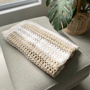 Puede incluir: Una manta de punto de crochet con rayas beige y blancas alternas. La manta está doblada y colocada sobre un otomano gris claro.