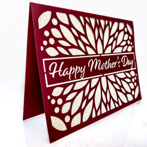 May include: A burgundy card with a white, intricate, laser-cut design of leaves and dots. The card has the text "Happy Mother's Day" in white lettering.