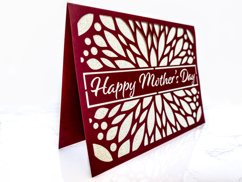 May include: A burgundy card with a white, intricate, laser-cut design of leaves and dots. The card has the text "Happy Mother's Day" in white lettering.