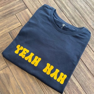 May include: A dark blue t-shirt with the phrase "Yeah Nah" printed in yellow bubble letters.