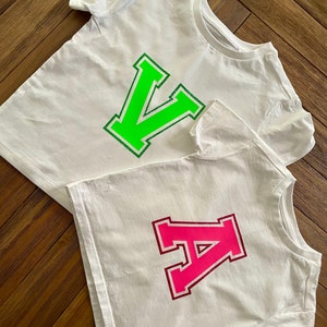 May include: Two white t-shirts with large letters on them. The top shirt has a green "V" and the bottom shirt has a pink "A".