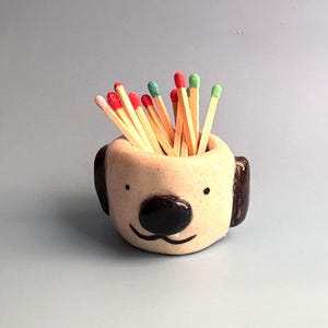 May include: A small, cream-coloured ceramic holder shaped like a dog's head, featuring dark brown ears and a black nose. The holder is filled with wooden matches that have red and green tips, creating a functional and decorative piece for storage.