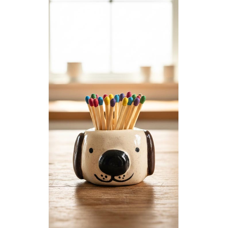 May include: A cream-coloured ceramic dog-shaped holder featuring dark brown ears, a black nose, and small black eyes. The holder is filled with a collection of wooden matches that have colourful heads in shades of red, blue, yellow, and green.