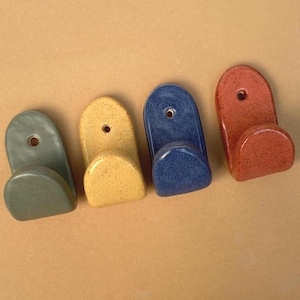May include: Four ceramic wall hooks in a row, each featuring a rounded top with a single mounting hole and a curved lower hook. The hooks are finished in solid, speckled glazes of sage green, mustard yellow, deep blue, and terracotta orange.