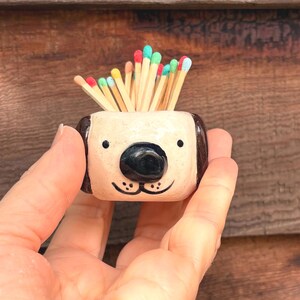 May include: A small, cream-coloured ceramic holder shaped like a dog's head, featuring dark brown ears, a black nose, and painted eyes and mouth. The holder is filled with wooden matches that have colourful tips in shades of red, green, blue, and yellow.
