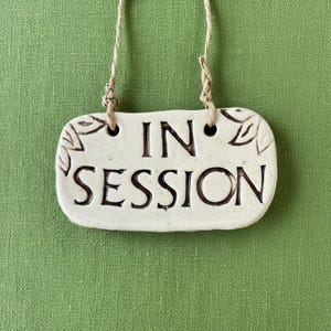 May include: Cream-colored ceramic sign with the words "IN SESSION" in dark brown lettering. The sign has a rectangular shape with rounded edges and leaf designs. It hangs from a natural twine string against a green background.