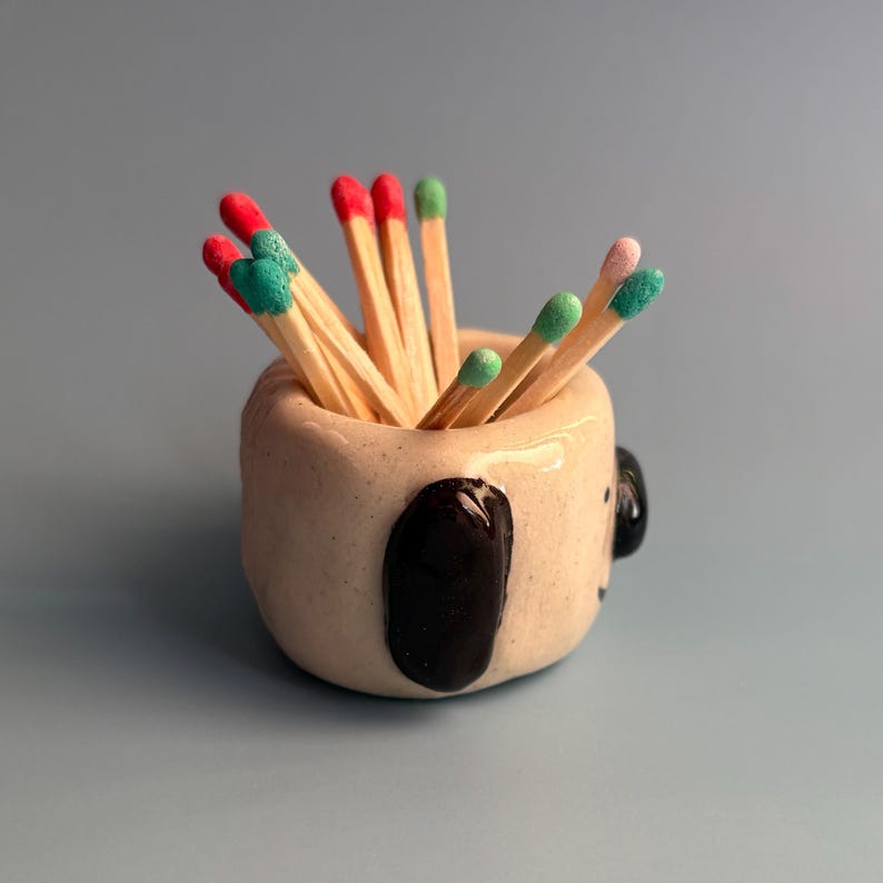 May include: A small, beige ceramic holder shaped like an animal, featuring a glossy black ear and a small black nose. The container is filled with wooden matches that have red, green, and light pink tips, creating a colourful display of matchsticks.