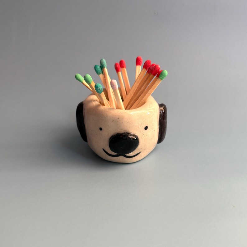 May include: A small, cream-coloured ceramic holder shaped like a dog's head with dark brown ears, a black nose, and two small eyes. The holder is filled with wooden matches that have red, green, and light pink tips, protruding from the top opening.