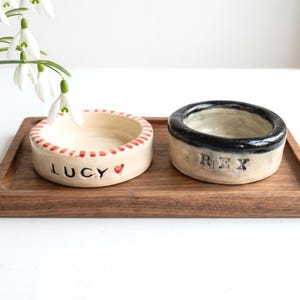 May include: Two round ceramic bowls on a rectangular wooden tray. The left bowl is cream-coloured with red stripes on the rim and the name LUCY with a red heart. The right bowl has a black rim and the name REX stamped in black letters.